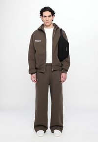 Rjav zip-up hoodie z belim grafičnim motivom, v kombinaciji z rjavimi širokimi hlačami in belimi čevlji. Črna torba na sling je nanešena čez ramo.