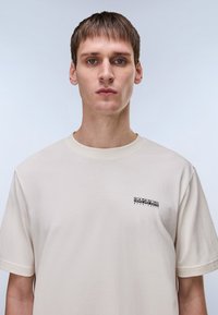 Βαμβακερό μπλουζάκι σε κρεμ απόχρωση με στρογγυλή λαιμόκοψη, κοντά μανίκια και μικρό μαύρο λογότυπο στην αριστερή πλευρά του στήθους. Απαλή υφή, άνετη εφαρμογή.