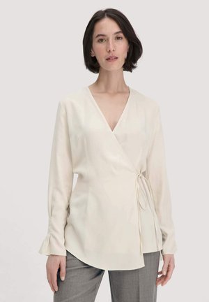 Vrouw met kort, donker haar in een crèmekleurige wikkelblouse met lange mouwen en grijze broek, staande tegen een lichte achtergrond.