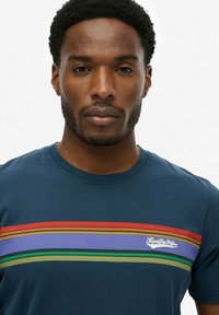 Camiseta de manga corta azul marino con rayas horizontales multicolores en rojo, amarillo, verde y púrpura; logo de la marca en el pecho.