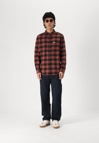 Camisa de franela de cuadros marrones y negros con un bolsillo en el pecho, diseño de botones, combinada con jeans oscuros y zapatillas blancas. Gafas de sol puestas.