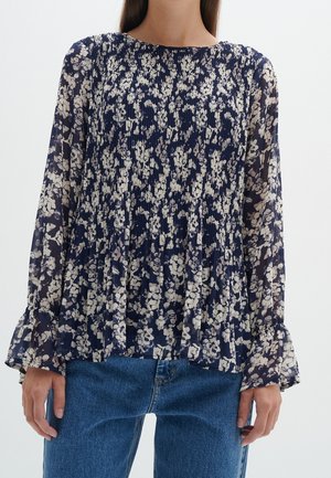 Mujer con blusa floral transparente azul marino de manga larga, puños con volantes y detalle plisado en el frente, combinada con jeans azules.