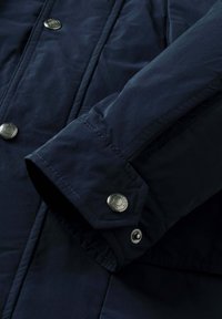 Navy blå jakkehæm med metalsnapknapper graveret "Hackett London" og synlige syningsdetaljer.