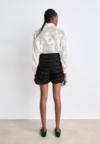 Blusa de encaje floral blanco con mangas largas, combinada con pantalones cortos negros texturizados con acentos metálicos, vista desde la espalda.