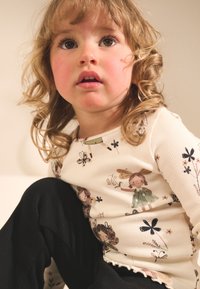 Haut crème à manches longues avec imprimés floraux et personnages ; texture côtelée. Bas noirs avec une finition lisse et des détails subtils.