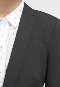 Černý blazer s jednou náprsní kapsou, nošený přes bílou košili s malým vzorovaným designem v červené a modré. Hladká textura.