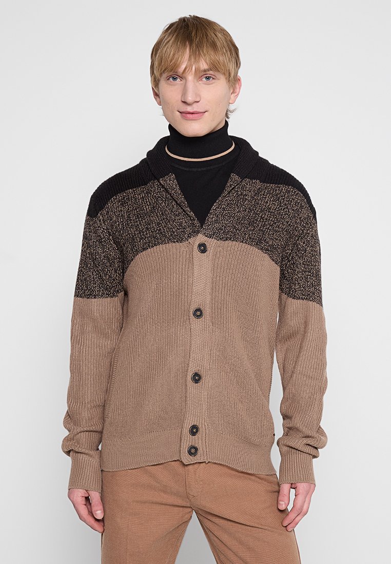 Brauner Strick-Cardigan mit strukturiertem Design, ausgestattet mit einem schwarzen Rippkragen und sieben Knöpfen an der Vorderseite, getragen über einem schwarzen Rollkragenpullover.