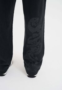 Pantaloni della tuta neri con una grafica lucida a forma di serpente lungo il lato, realizzati in un tessuto morbido con un fit rilassato e polsini affusolati.