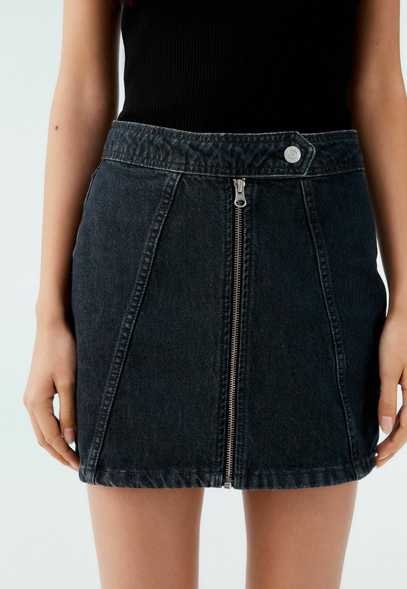 Svart denim mini kjol med centrerad dragkedja, figursytt midjeband och diagonala sömmar. Tyget har en lätt textur. Knappdetalj vid midjan.