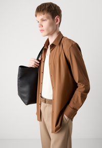 Camicia marrone a maniche lunghe in materiale leggero, indossata sopra una canotta bianca, abbinata a pantaloni beige e una borsa tote in pelle nera.