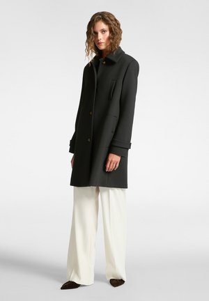 Cappotto nero con un colletto alto, bottoni frontali, tasche laterali e una silhouette strutturata; indossato sopra pantaloni bianchi a gamba larga.