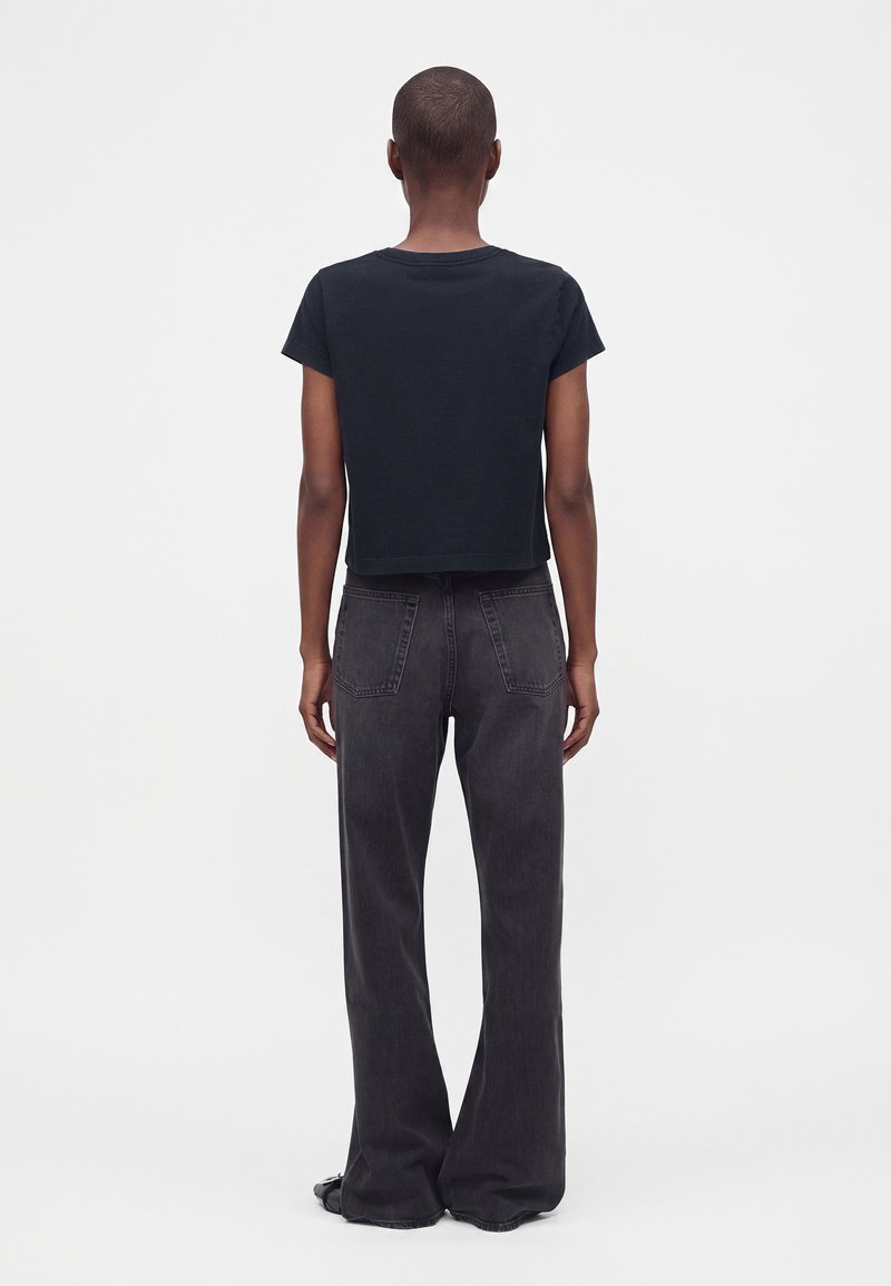 Personne portant un t-shirt noir court à manches courtes et un jean ample gris foncé taille haute, debout de dos sur un fond uni clair.