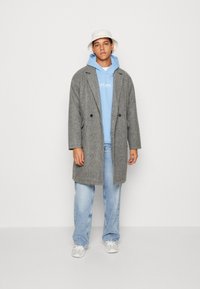 Cappotto di lana grigio con due bottoni frontali, indossato sopra un hoodie azzurro chiaro, abbinato a jeans di denim chiaro e un cappello a secchiello bianco.