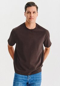 Mężczyzna z krótkimi ciemnymi włosami, ubrany w gładką ciemno-brązową koszulkę i niebieskie dżinsy, stoi z rękami za plecami.