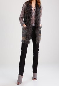 Donna con una blusa a stampa leopardata, un lungo gilet di pelliccia grigia testurizzata, pantaloni neri e stivaletti grigi con la punta aperta, in piedi con le mani nelle tasche.