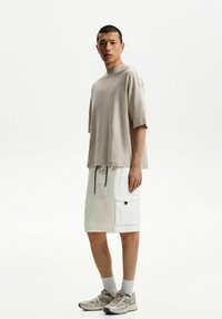 Béžové oversized tričko s kleslými rameny, spárované s bílými šortkami s bočními kapsami. Tenisky s světle šedými a béžovými akcenty.