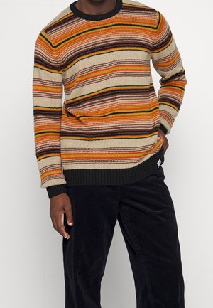 Man wearing a striped knit sweater in orange, yellow, beige, and brown tones paired with black corduroy pants.