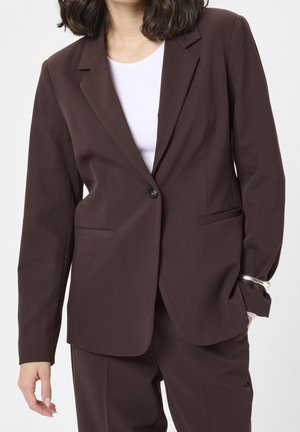 Frau trägt einen dunkelbraunen, maßgeschneiderten Blazer und passende Hose mit weißem Hemd darunter, steht mit einer Hand in der Tasche.