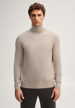 Junger Mann mit lockigem Haar, der einen hellbeigen Rollkragenpullover und dunkle Hose trägt, steht vor einem einfarbigen Hintergrund.