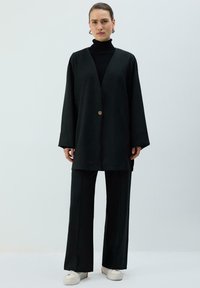 Melns garš bleizeris ar V veida izgriezumu, pārmērīgi lielām piedurknēm un vienu pogu aizdari, kopā ar saskaņotām platu kāju biksēm un baltām sporta kurpēm.