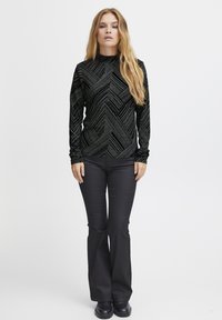 Schwarzes Langarmoberteil mit geometrischem Muster, aus einem strukturierten Stoff gefertigt. Kombiniert mit figurbetonten schwarzen Hosen und flachen schwarzen Schuhen.