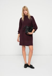 Abito lavorato a maglia bordeaux con scollo alto e maniche a palloncino, abbinato a una borsa a tracolla nera con coulisse e scarpe nere slip-on.