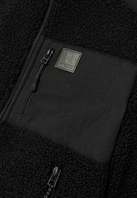 Zwarte fleecjacket met een textuur van een pluizige stof en een glad paneel op de borst. Inclusief een ritszak en een merkpatch.