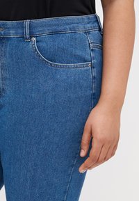 Close-up of a person wearing blue denim jeans and a black top, with their right hand resting by their side.