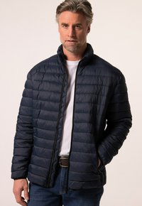 Námořnická péřová bunda s pro šívaným designem, vysokým límcem, zipovým uzávěrem a bočními kapsami. Nositá přes bílé tričko a v kombinaci s modrými džíny.