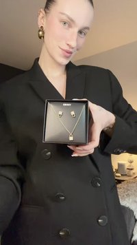 Set de bijuterii cu nuanță aurie, format dintr-un colier cu pandantiv în formă de lacăt și cercei asortați, expus într-o cutie neagră DKNY. Se observă accente pe suprafața texturată.