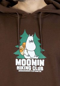 Ruskea huppari, jossa on piirroselokuvahahmo kantamassa reppua, taustalla vihreitä puita, ja teksti "MOOMIN HIKING CLUB" paksulla valkoisella fontilla.