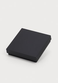 Négyzet alakú, fekete, zárt doboz, a tetjén diszkrét, dombornyomott "MAPLE" felirattal, egy sima fehér felületen elhelyezve.