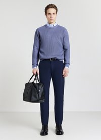 Mann in hellblauem Hemd, lila Strickpullover, marineblauen Trouser und schwarzen Stiefeln, der in seiner rechten Hand eine große schwarze Ledertasche hält.