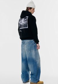 Svart hoodie med grafiskt tryck på baksidan, matchad med lösa blå denimjeans och beige skor. Accentuerande mössa ger en extra textur.