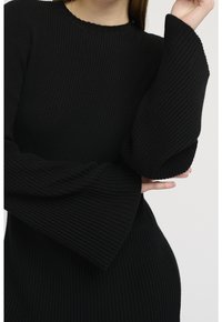 Svart ribbad klänning med rund halsringning och utsvängda, strukturerade ärmar. Tyget har en mjuk, stickad yta som framhäver en figurnära silhuett.