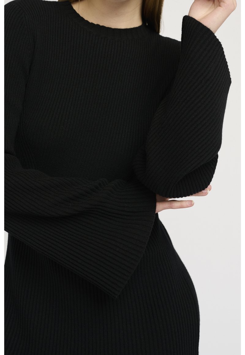 Svart ribbad klänning med rund halsringning och utsvängda, strukturerade ärmar. Tyget har en mjuk, stickad yta som framhäver en figurnära silhuett.