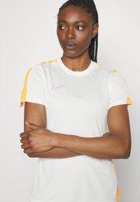 Valge spordipaita, millel on oranžid aksendid õlgadel, ümmargune kaelus, lühikesed varrukad ja rindkere peal väike lilla Nike logo.