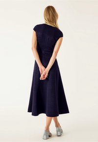 Mörkblå midi-klänning med korta ärmar, figurnära liv och utsvängd kjol. Texturerad tyg med synliga sömmar, tillsammans med grå blockklackar.