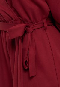 Κοντινό πλάνο δέσμου υφασμάτινης ζώνης δεμένης σε φιόγκο πάνω σε ένδυμα βαθύ κόκκινου χρώματος με λεπτομέρεια σούρας στη μέση.