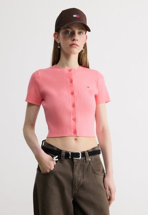 Young person wearing a brown cap, pink short-sleeve ribbed buttoned crop top, and brown high-waisted pants with a black belt.