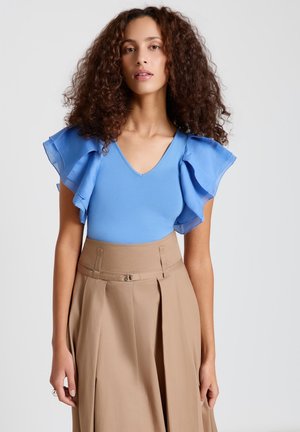 Femme aux cheveux bouclés portant un haut bleu à manches courtes à volants, rentré dans un pantalon plissé beige taille haute avec un détail de ceinture.