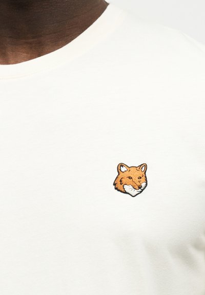 Κοντινό πλάνο ενός ανοιχτό μπεζ T-shirt που δείχνει μια μικρή κεντημένη πορτοκαλί αλεπού στο αριστερό μέρος του στήθους.