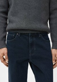 Jeans de mezclilla azul marino con un corte clásico, que cuentan con bolsillos delanteros y cierre de cremallera. Combinados con un suéter gris de canalé.