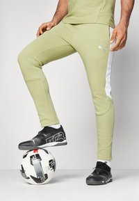 Pantalons de sport Puma vert clair avec des bandes blanches sur les côtés, associés à des chaussures de sport noires, reposant sur un ballon de soccer noir et blanc.