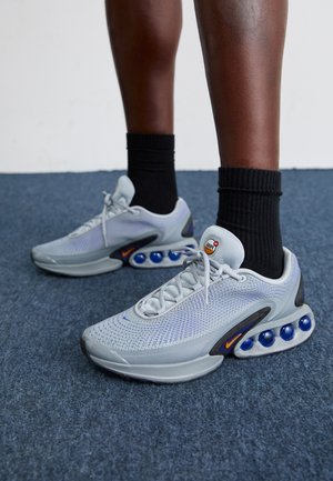 Helle graue Sportsneaker mit Mesh-Obermaterial, strukturiertem Muster, blauen Luftpolster-Gegenständen und schwarzen Schnürsenkeln. Getragen mit schwarzen Crew-Socken.