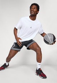 Hvid T-shirt med sort logo, sorte shorts med hvide striber og røde og sorte sportsko. Holder en struktureret basketball.