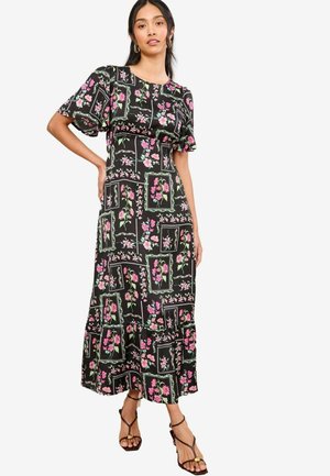 Robe maxi fleurie noire à manches courtes volantées, présentant un motif de fleurs roses et vertes disposées en grille, avec un ourlet à volants.