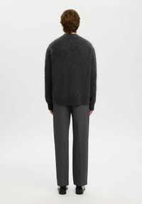 Homme debout de dos portant un pull sombre et pelucheux, un pantalon gris ajusté et des chaussures noires sur un fond blanc uni.