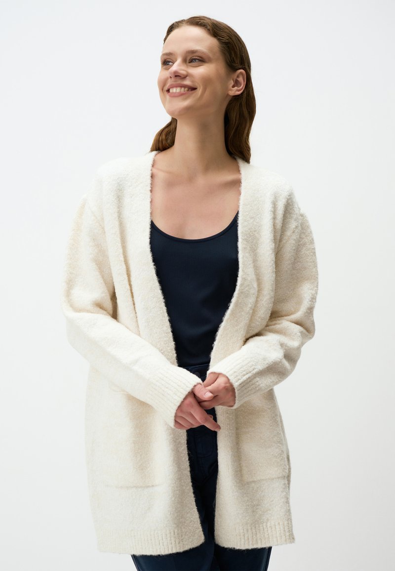 Cardigan bianco e morbido con maniche lunghe, apertura frontale e polsini a coste. Indossato sopra un top scuro, con tasche laterali e texture morbida.