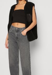 Gilet in pile nero senza maniche con tasca con zip, abbinato a jeans grigi a vita alta eamp; larghi con tasche frontali e chiusura a bottone.